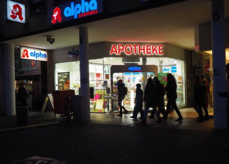 Foto de Personas caminando frente a una farmacia iluminada Alpha Apotheke por la noche en Darmstadt, Alemania - Imagen libre de derechos