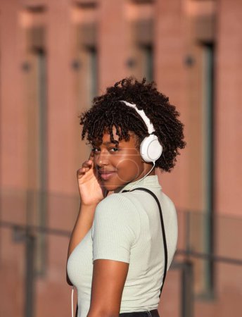 Téléchargez les photos : Une jolie adolescente latino-américaine aux cheveux bouclés écoutant de la musique avec des écouteurs blancs dans la rue - en image libre de droit
