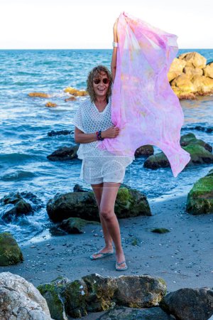 Photo for A vertical of Hispanic female model over 50 years with pink scarf and sunglasses on the beach - Royalty Free Image