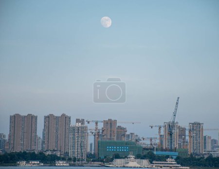 Photo for The white full moon in blur sky over a skyline with construction cranes at an early morning - Royalty Free Image