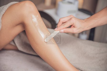 Photo for A therapist applying warm wax with a wooden spatula on the client's leg. - Royalty Free Image