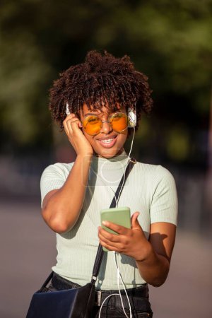 Téléchargez les photos : Une jeune femme latino-américaine avec des cheveux afro et des lunettes souriant joyeusement en utilisant smartphone et écouteurs - en image libre de droit
