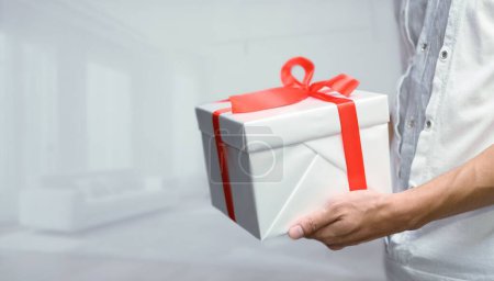 Foto de Un enfoque selectivo de un hombre sosteniendo un regalo con un arco, en una habitación blanca, el concepto de dar regalos para Año Nuevo, cumpleaños y otras ocasiones - Imagen libre de derechos