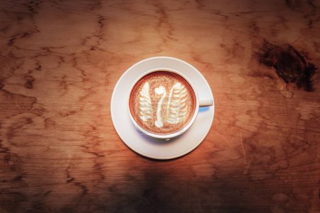 Foto de Una vista superior de una taza de café con leche decorada con una espuma en forma de ganso en una mesa de madera - Imagen libre de derechos