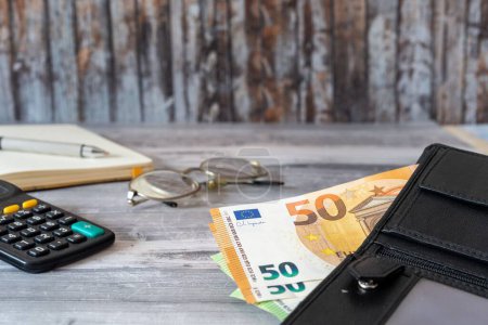 Téléchargez les photos : Les billets en euros dans un portefeuille, une calculatrice, des lunettes sur une table - en image libre de droit