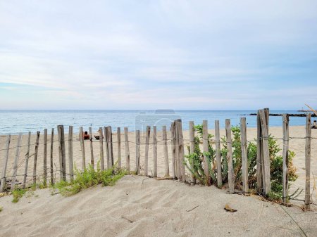 Téléchargez les photos : Une plage de sable avec une clôture en bois capturée contre le paysage marin pittoresque sous le ciel clair - en image libre de droit