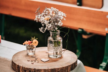 Foto de Un primer plano de flores en un jarrón de vidrio y anillos de boda en un plato de madera - Imagen libre de derechos
