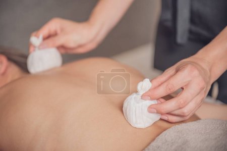 Photo for A closeup shot of the masseur's hands massaging her client's back with Thai herbal compress balls. - Royalty Free Image