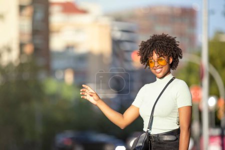 Photo for A beautiful afro-haired young woman standing on the street and taking a taxi with her hand up - Royalty Free Image