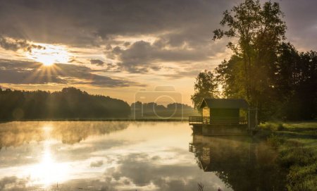 Téléchargez les photos : Un étang pittoresque avec une cabane solitaire sur le rivage et un magnifique coucher de soleil en arrière-plan - en image libre de droit