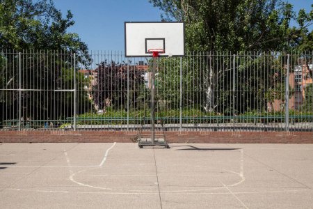 Photo for An outdoor fenced basketball court, with a backboard, surrounded by green trees in a sunny day - Royalty Free Image