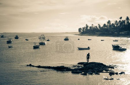 Foto de Una escala de grises de la silueta de la persona de pie en una isla rocosa con barcos en el fondo - Imagen libre de derechos