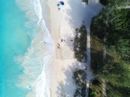 An aerial top view of the blue ocean with people on a sandy beach near the trees in Waimanalo, Oahu, Hawaii