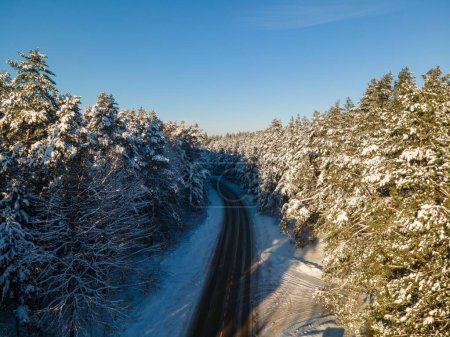 Téléchargez les photos : Un drone d'une route asphaltée traversant une forêt luxuriante et enneigée sous le ciel bleu - en image libre de droit