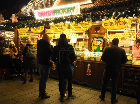 Foto de Una vista nocturna de la fábrica de dulces, un puesto de confitería en el mercado de Navidad y gente parada frente a ella en Alemania - Imagen libre de derechos