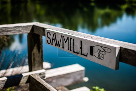 Foto de Un primer plano del letrero con una escritura "aserradero" y una señal de mano que indica la dirección en un lago y los árboles de fondo - Imagen libre de derechos