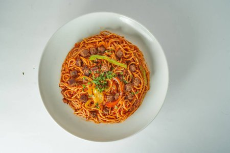 Foto de Una vista superior de los espaguetis con salsa de tomate, rebanadas de carne y pimientos de colores en rodajas en un plato blanco en una mesa blanca - Imagen libre de derechos