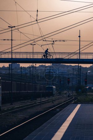 Téléchargez les photos : Une silhouette de cycliste montant sur un pont au-dessus du chemin de fer contre le coucher du soleil - en image libre de droit