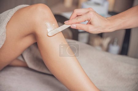 Téléchargez les photos : Un thérapeute applique de la cire chaude avec une spatule en bois sur la jambe du client. - en image libre de droit