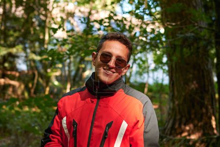 Téléchargez les photos : Une mise au point peu profonde de beau jeune homme hispanique avec des lunettes de soleil et manteau posant dans la nature - en image libre de droit
