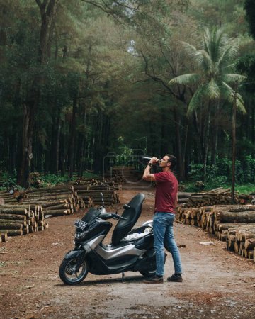 Téléchargez les photos : Une verticale d'un modèle masculin d'eau potable près d'un scooter dans une forêt indonésienne à Java - en image libre de droit