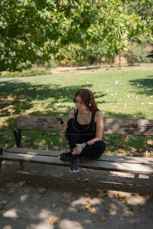 A young redhead girl, looking at the smartphone, with music headphones sitting on a park bench
