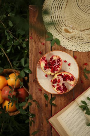 Photo for A vertical top view shot of a sliced pomegranate on a wooden table with a hat, book and fruit basket. - Royalty Free Image