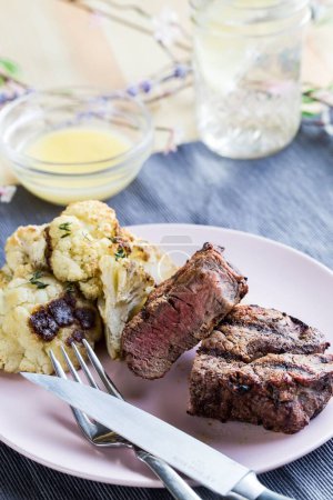 Photo for A vertical closeup shot of the steak dinner on a white plate with a sauce and a glass of water on a blue fabric - Royalty Free Image