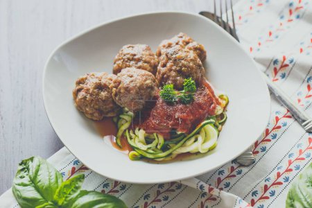 Photo for A closeup of the meatballs with tomato sauce and zoodles on a white plate next to a fork and spoon - Royalty Free Image