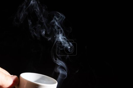 Téléchargez les photos : Une main tenant une tasse blanche avec de la fumée sur un fond noir - en image libre de droit