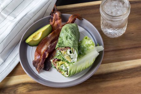 Photo for A closeup of the lettuce wraps with bacon and avocado on a gray plate with a glass of iced water on a wooden table - Royalty Free Image