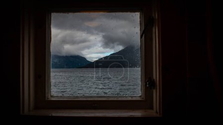 Foto de Una vista panorámica del mar con montañas bajo el cielo sombrío visto desde una pequeña ventana - Imagen libre de derechos