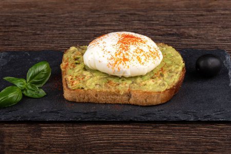 Photo for An avocado toast with poached egg, olive, and basil leaves on a stone board for serving - Royalty Free Image