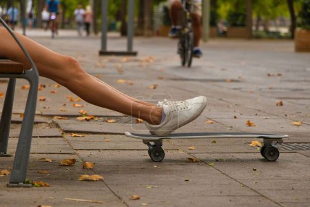 Photo for A girl sitting on a bench in the pedestrian street and putting her feet on the skateboard - Royalty Free Image