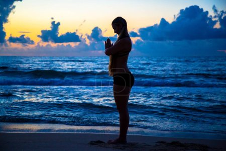 Téléchargez les photos : Une femme caucasienne sur la plage avec la pose de prière (Pranamasana) de Surya Namaskar, au coucher du soleil - en image libre de droit
