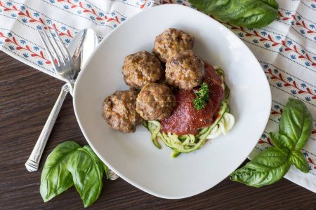 Photo for A top view of the meatballs with tomato sauce and zoodles on a white plate next to a fork and spoon on a wooden table - Royalty Free Image