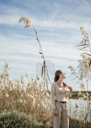 Foto de Una foto vertical de una joven y elegante hembra posando cerca de juncos altos creciendo en la orilla de un lago - Imagen libre de derechos