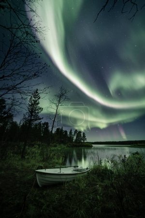 Photo pour Un plan vertical des aurores polaires dans le ciel étoilé au-dessus du lac avec un bateau sur un rivage - image libre de droit