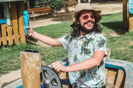 Foto de Un hombre caucásico feliz con el pelo largo y una barba sosteniendo un cono de helado azul en un parque infantil - Imagen libre de derechos