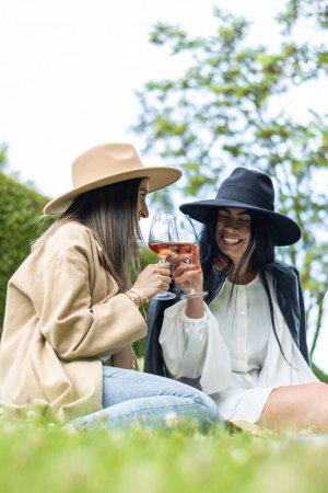 Photo for A vertical shot of two women friends in hats sitting on the grass and toasting glasses of wine - Royalty Free Image