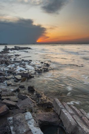 Téléchargez les photos : Vue verticale à longue exposition des vagues de mer atteignant les pierres côtières au coucher du soleil - en image libre de droit