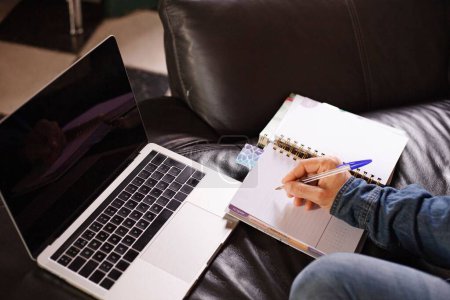 Foto de Un primer plano de mano masculina con bolígrafo anotando notas en un cuaderno y utilizando un ordenador portátil en el sofá en casa - Imagen libre de derechos