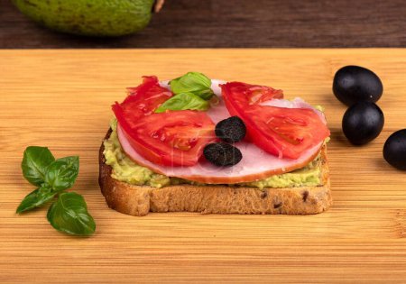 Foto de Un sándwich con aguacate, rodajas de jamón y tomate con aceitunas en una tabla de cortar de madera. - Imagen libre de derechos
