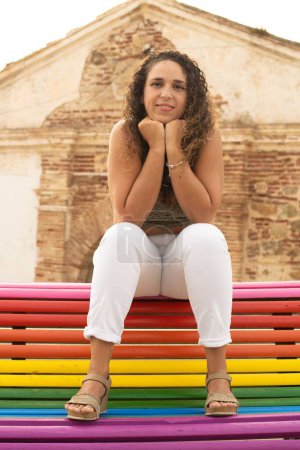 Foto de Joven morena en jeans blancos sentada en un banco con los colores de la bandera LGBT - Imagen libre de derechos