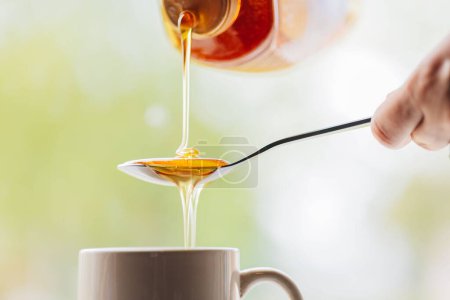 Foto de Un primer plano de una persona vertiendo miel natural en una cuchara para poner en el té de la mañana - Imagen libre de derechos