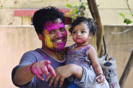 Photo for The Indian man holding a baby girl and smiling while pointing at the camera both covered in colorful paints - Royalty Free Image