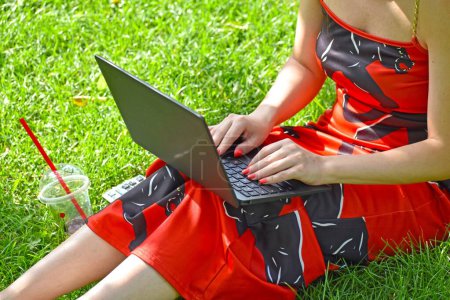 Foto de Un primer plano de una chica que trabaja en su computadora portátil mientras está sentada en el césped en un parque con un vestido de verano - Imagen libre de derechos