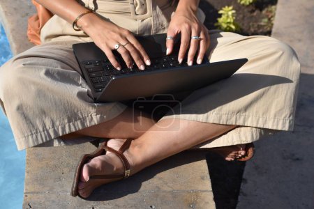 Photo for A woman sitting on a ledge and using a laptop at the sunlight - Royalty Free Image