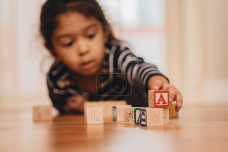 Foto de Un enfoque selectivo de bloques de letras de madera, con una niña preescolar en el fondo, acostada en el suelo en casa, el concepto de educación temprana - Imagen libre de derechos