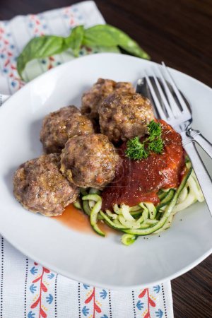Photo for A vertical closeup shot of the meatballs with tomato sauce and zoodles on a white plate with a fork and spoon on a wooden table - Royalty Free Image